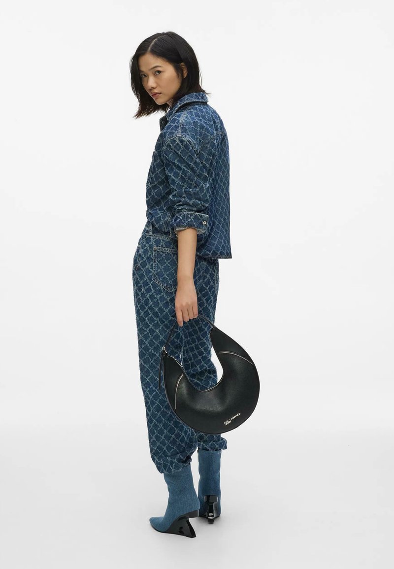 Femme vêtue d'une tenue en jean bleue à motifs, tenant un sac à main noir en forme de croissant, portant des bottines bleues à talons, debout devant un fond blanc.