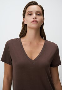 T-shirt marrone a V con maniche corte, texture liscia, forma aderente e dettagli di cucitura sottili. Nessun motivo o accento visibile.