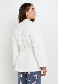 Rospetto bianco e texturizzato con maniche larghe e vita legata, indossato sopra pantaloni da pigiama a fantasia blu con disegni di uccelli rosa.