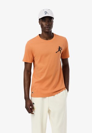 Hombre con gorra de béisbol blanca, camiseta naranja con un pequeño gráfico negro en el pecho y pantalones blancos con logos bordados, posando frente a un fondo blanco.