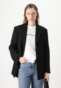 Svart skreddersydd blazer med to knapper, spiss lapel og sidelommer, lagd over en hvit t-skjorte med grafikk og lyseblå denimjeans.