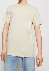 Camiseta de algodón beige de manga corta, con cuello redondo y un logo blanco "CK" en el lado izquierdo del pecho, combinada con jeans de color azul claro.