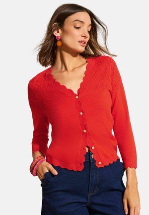 Žena v červenom kardigáne s oblými okrajmi a textúrovaným pleteným živôtikom, v kombinácii s modrými džínsami, oranžovými náušnicami a ružovými náramkami.
