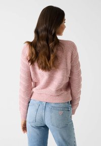 Pull en maille rose clair avec un motif texturé, poignets et ourlet côtelés, associé à un jean bleu clair avec un écusson triangulaire.