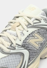 Witte en grijze New Balance hardloopschoen met beige veters, mesh zijkanten, en het "N" logo van het merk op het zijpaneel.