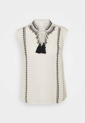 Mouwloze crèmekleurige blouse met zwarte geometrische borduursels langs de voorkant en kraag, voorzien van een strikkraag met twee zwarte kwastjes.