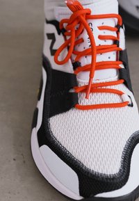 Hvide mesh sneakers med sorte accenter og lyse orange snørebånd. Har en tekstureret overflade og forstærket hæl. Tæt på udsigt.