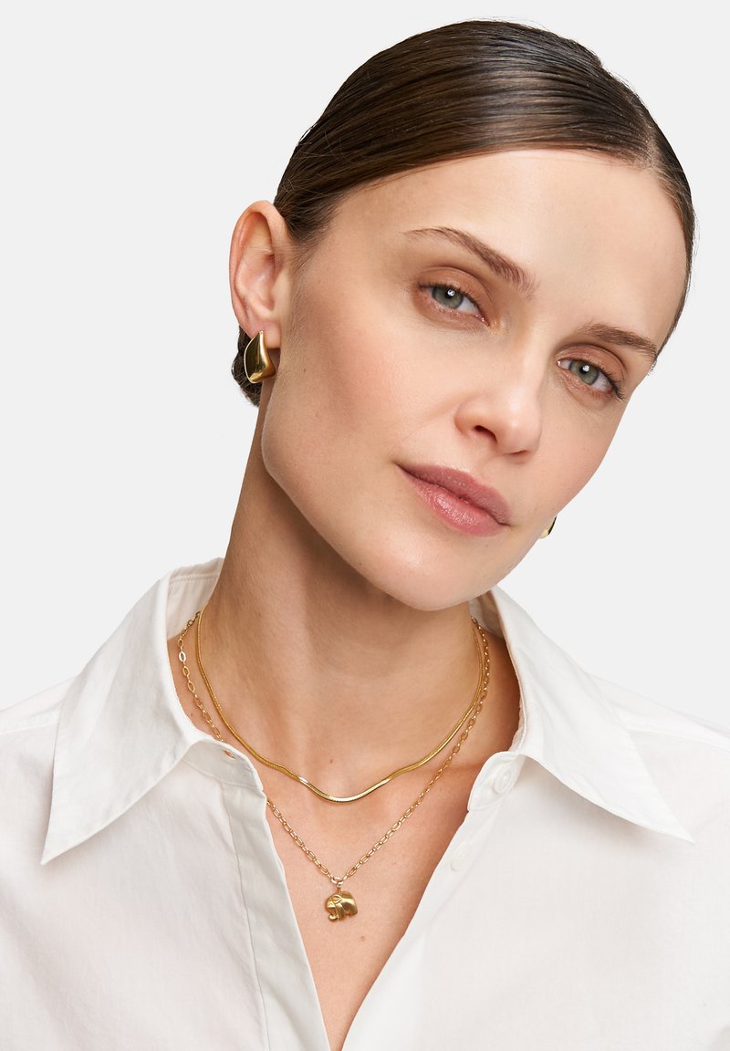 Goldschmuck mit geschichteten Halsketten, einer gebogenen Kette, einem Anhänger und auffälligen goldenen Ohrringen, kombiniert mit einem weißen Hemd.