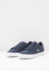 Paire de baskets bleu marine à lacets avec semelles blanches et petit logo crocodile vert sur le côté extérieur.