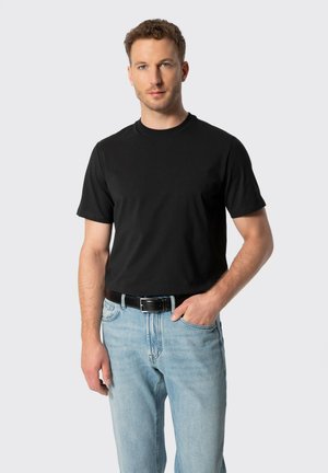 Joven con el pelo rizado y corto, lleva una camiseta negra lisa y vaqueros azul claro con un cinturón negro, de pie con una mano en el bolsillo.