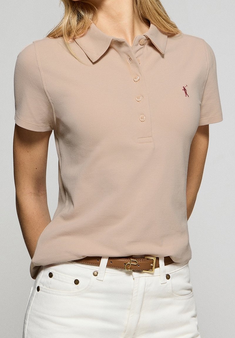 Femme portant un polo beige à manches courtes avec des boutons et un pantalon blanc avec une ceinture marron, debout les mains dans le dos.
