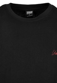 T-shirt Urban Classics noir à col rond avec un petit logo rouge en écriture sur la poitrine et une étiquette de taille visible à l'intérieur du col.