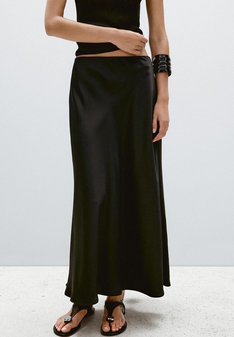 Black maxi skirt in smooth, lightweight fabric, featuring a fitted waistband and flowing silhouette. Paired with black sandals and an arm cuff.