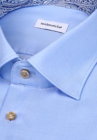 Chemise habillée bleu clair en tissu lisse et texturé, avec un col boutonné et un intérieur à motif cachemire. Étiquette visible.