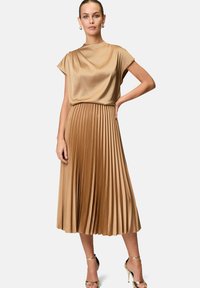 Satin geplooide jurk in een warme beige kleur met een gedrapeerd bovenstuk en een broeklang rok; gecombineerd met metallic hakken.