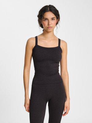 Woman with dark hair in braided updo wearing a fitted black sleeveless top and matching high-waisted pants, standing against a white background.