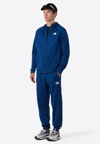 Blauwe trainingsoutfit van lichtgewicht materiaal, met een rits hoodie en elastische manchetten, gecombineerd met zwart-witte sneakers.