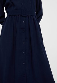 Abito camicia blu navy con maniche lunghe, vita con cintura, chiusura frontale con bottoni e pieghe per una silhouette fluida.