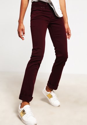 Persoon draagt slank zittende donker bordeauxrode broek met opgerolde pijpen en witte sneakers met gouden accenten, staand tegen een effen achtergrond.