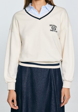 Mujer que lleva un suéter crema con cuello en V, ribete negro y logo en el pecho, sobre una camisa azul con cuello, combinado con una falda de mezclilla oscura.