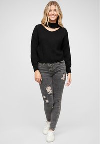 Fekete kötött pulóver magas nyakkal és kivágott részletekkel, szürke, tépett skinny farmerral és fehér sportcipővel párosítva.