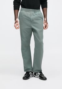 Svijetlozelene hlače od tkanine s elastičnim pojasom, ravnog kroja i suptilne teksture, uparene s crnim sportskim tenisicama koje imaju srebrne detalje.