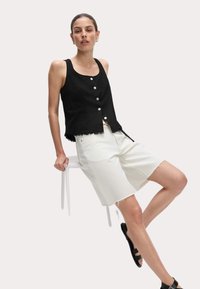 Vrouw zittend op de rand van een witte kruk, gekleed in een zwarte mouwloze blouse met knopen, witte knie-lange shorts en zwarte sandalen.