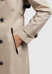 Beige regnkappa med en slät, matta yta, prydd med svarta knappar och en dragkedjeficka. Manschetten har räfflad detaljering för textur.