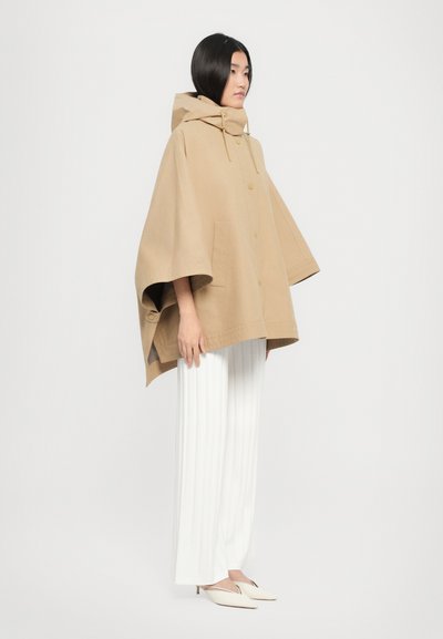 Beige huppari poncho, joka on valmistettu pehmeästä kankaasta ja on löysästi istuva, sisältäen suuret sivutaskut ja napit. Yhdistettynä valkoisiin laskostettuihin housuihin.