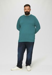 Türkisfarbener Pullover mit langen Ärmeln und rundem Ausschnitt; kombiniert mit dunklen Jeans und weißen Turnschuhen. Das Modell posiert mit den Händen hinter dem Rücken.