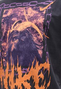 Σκούρα μωβ και πορτοκαλί γραφικό κεφάλι κατσίκας με κέρατα μέσα σε μωβ πλαίσιο αλυσίδας με πορτοκαλί σχήματα σαν φλόγες στο κάτω μέρος.