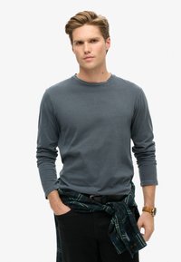 Grijze lange mouw shirt van katoen, met een ronde halslijn. Gepaard met donkere broek en een groen geruite flanellen stof om de taille geknoopt.