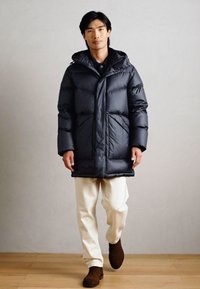 Casaco acolchoado em cinza escuro, com capuz, fecho com zip e bolsos frontais. Usado sobre calças em tom creme e botas castanhas, num chão de madeira.