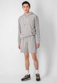 Sweat à capuche gris avec poche, logo blanc et rayures sur les manches, associé à un short assorti. Porté avec des chaussures de sport multicolores.
