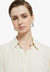 Camicia beige chiaro con colletto, caratterizzata da una chiusura frontale con bottoni. Il modello indossa orecchini bianchi testurizzati con elementi circolari.