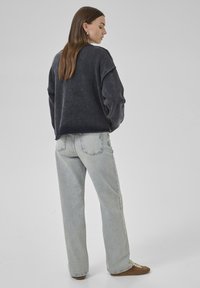 A person with long brown hair, wearing a dark sweater and light jeans, stands with their back to the camera against a neutral background.