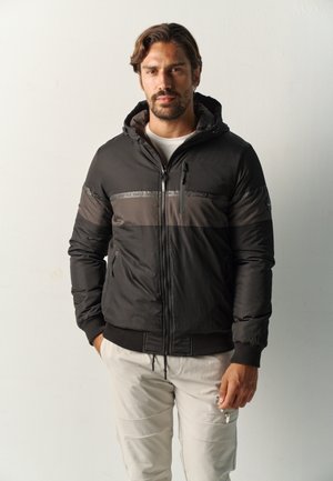Uomo con capelli corti e scuri e barba, che indossa una giacca con cappuccio nera e grigia e pantaloni beige chiari, in piedi davanti a una parete bianca semplice.