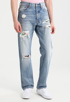 Hombre con vaqueros rotos de color azul claro con parches visibles de tela floral bajo los desgarros, combinado con zapatillas blancas y una camisa rosa.