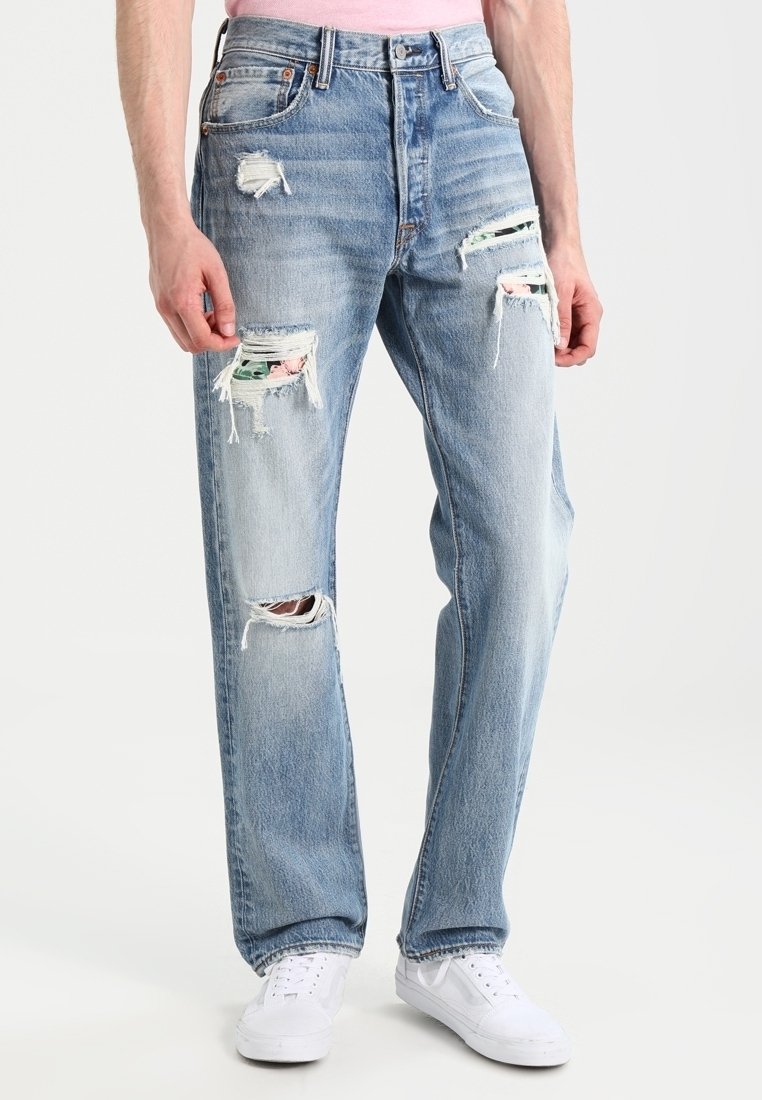 Homme portant un jean déchiré bleu clair avec des pièces de tissu à motif floral visibles sous les déchirures, associé à des baskets blanches et une chemise rose.