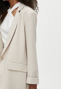 Blazer beige avec un seul bouton, des revers et des manches retroussées. Tissu lisse et coupe cintrée. Détail subtil de poche sur la gauche.