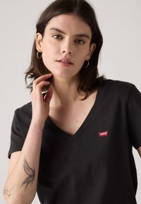 Juoda medvilninė V formos kaklo marškinėliai trumpomis rankovėmis su raudona Levi’s logotipo ženklelio detalė. Matoma modelio tatuiruotė ant rankos. Vienoda fono spalva.