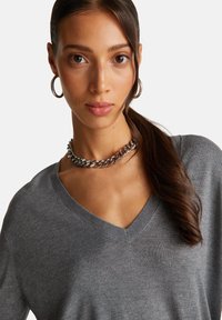 Grauer V-Ausschnitt-Pullover aus weichem Material, mit geripptem Halsausschnitt und subtiler Struktur, kombiniert mit einer silbernen Kettenhalskette und Creolen.