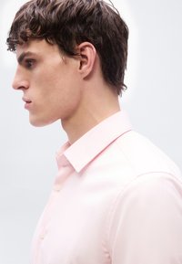 Νεαρός άνδρας με κοντά, καφέ σγουρά μαλλιά που φοράει ένα ανοιχτό ροζ πουκάμισο με κουμπιά, στρέφεται προς τα αριστερά μπροστά σε ένα απλό φωτεινό φόντο.