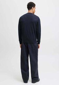 Uomo in piedi con le spalle rivolte verso di noi, che indossa un maglione blu navy e pantaloni scuri larghi, su uno sfondo chiaro e uniforme.