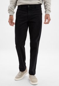 Pantalons en coton noirs à coupe droite, avec fermeture à bouton et design à cinq poches. Portés avec des baskets de couleur claire.