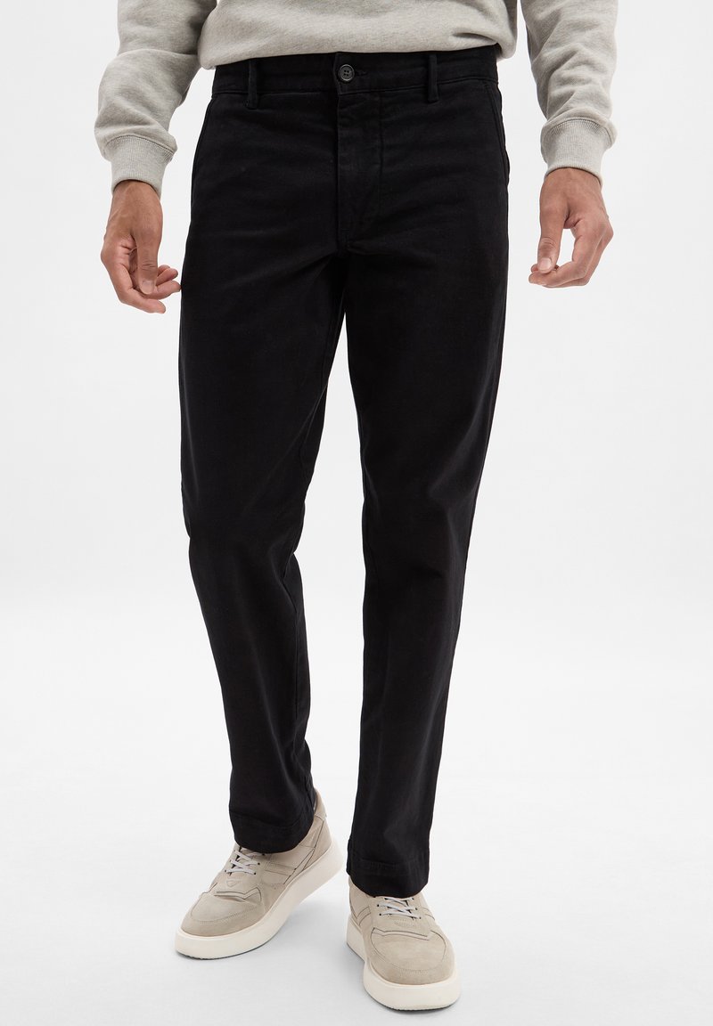 Pantalons en coton noirs à coupe droite, avec fermeture à bouton et design à cinq poches. Portés avec des baskets de couleur claire.