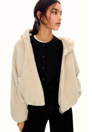 Korte faux fur jas in lichtbeige, met een capuchon, ritssluiting en voorzakken. Draagt over een zwarte geknoopte bovenkleding.