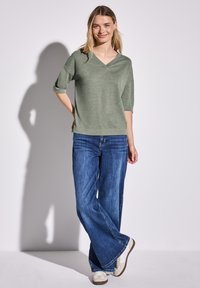 Luźno dopasowany, zielony top z dekoltem w serek, wykonany z miękkiego materiału, w połączeniu z szerokimi niebieskimi dżinsami i białymi tenisówkami, prezentujący swobodny styl.