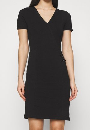 Kobieta ubrana w czarną sukienkę z krótkim rękawem, z dekoltem w kształcie litery V i dwoma ozdobnymi srebrnymi klamrami na prawej talii.