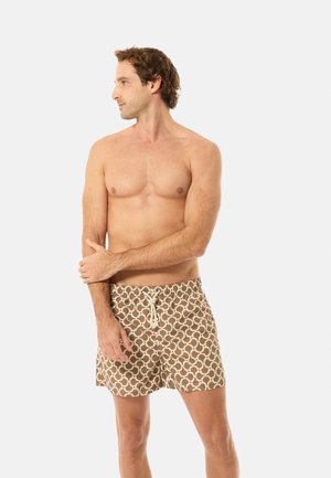Oberkörperfreier Mann, der nach links schaut, trägt beige Badeshorts mit einem weißen und roten geometrischen Muster und einem weißen Kordelzug.